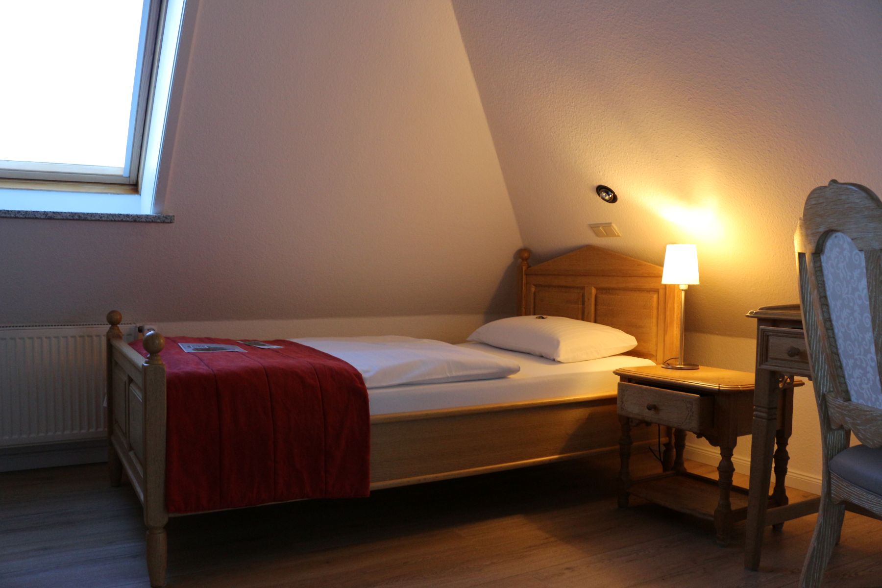 Cozy single room with wooden bed, bedside table and lamp under a sloping roof, warmly lit