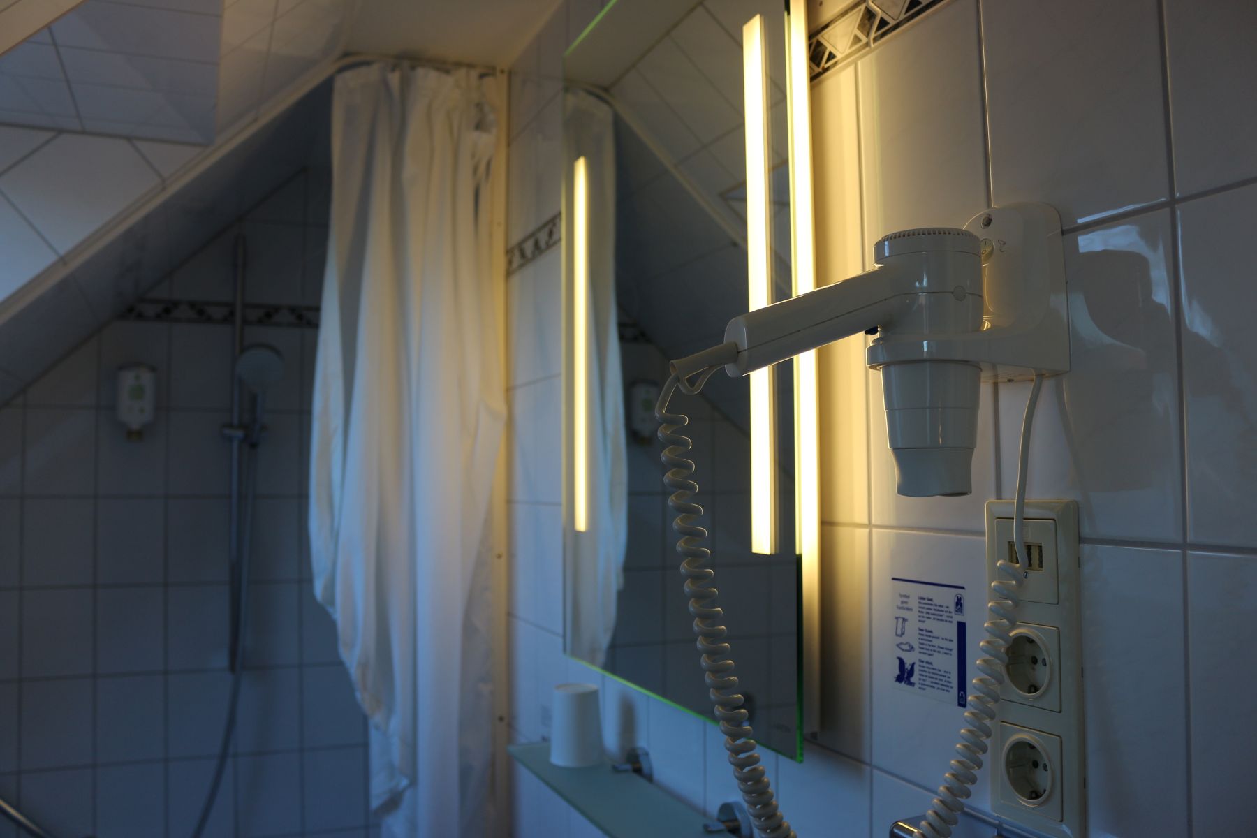 Bathroom with illuminated mirror, wall-mounted hairdryer and view of a bathtub with shower curtain under a sloping roof