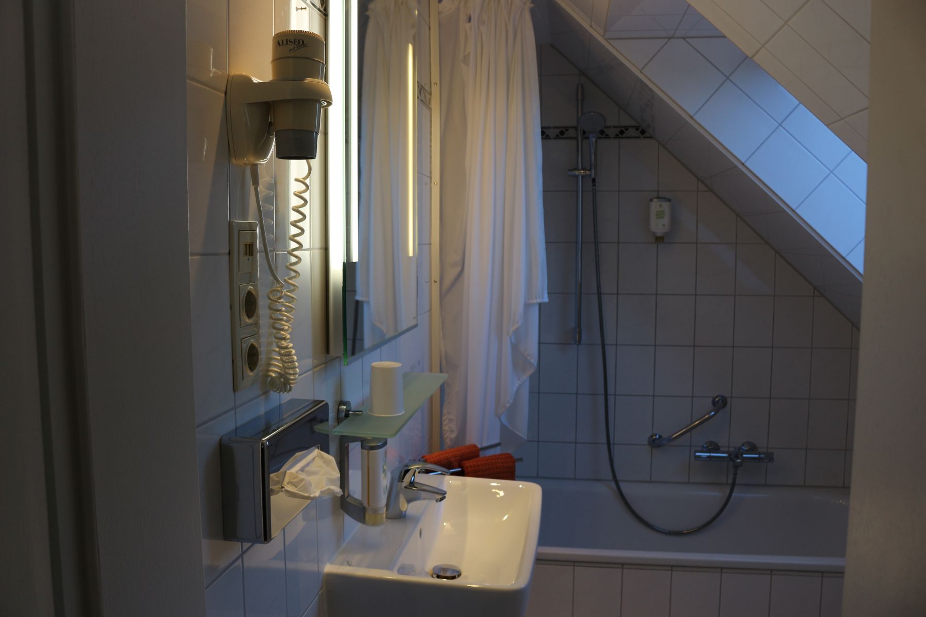 Small bathroom under a sloping roof with washbasin, mirror lighting, wall-mounted hairdryer and bathtub with shower curtain